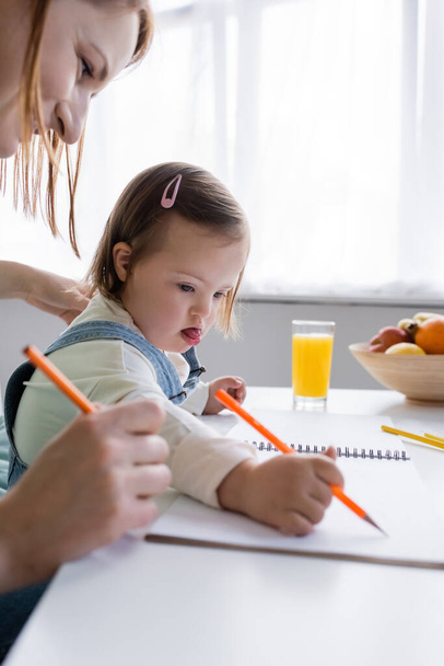 Bambino con sindrome di Down disegno su carta vicino a madre e succo d'arancia in cucina  - Foto, immagini