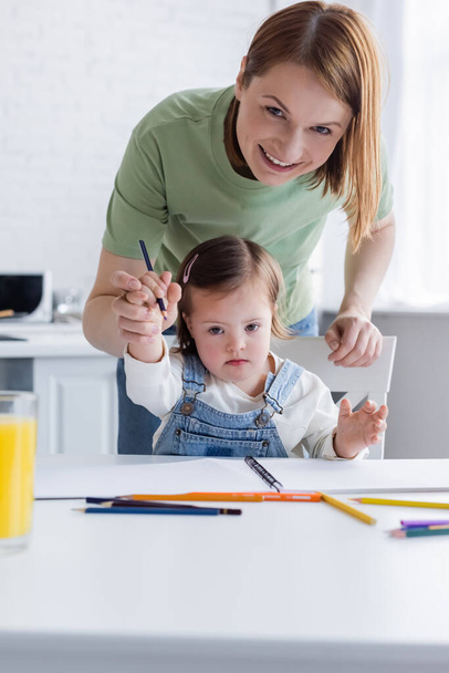 Smiling woman looking at camera near child with down syndrome and color pencils  - Φωτογραφία, εικόνα