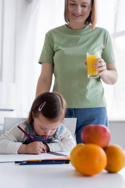 Kid met down syndrome tekening in de buurt van glimlachende moeder met sinaasappelsap in de keuken  - Foto, afbeelding