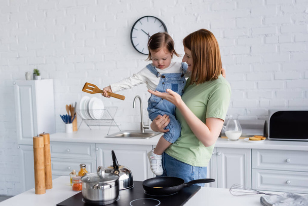 Smiling woman holding daughter with down syndrome holding spatula near stove in kitchen  - 写真・画像