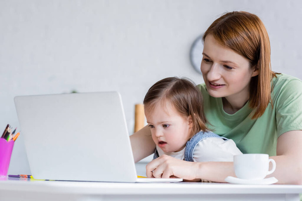Lächelnde Freiberuflerin mit Laptop neben Tochter mit Down-Syndrom und Kaffee zu Hause  - Foto, Bild