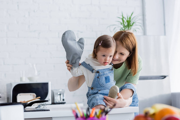 Улыбающаяся женщина стоит рядом с ребенком с синдромом Дауна и выпечкой перчатки на кухне  - Фото, изображение