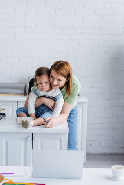 Smiling woman pointing at laptop near daughter with down syndrome in kitchen  - Fotografie, Obrázek