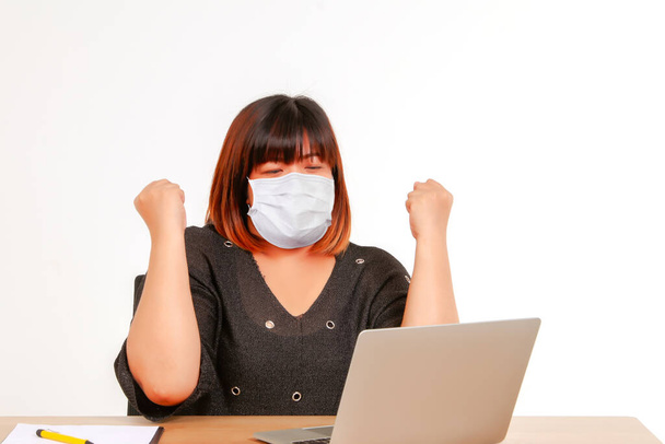 Trabajo conceptual en casa: Mujer asiática gorda con una máscara protectora contra el coronavirus (covid-19), sentada en un escritorio, usando una computadora portátil, expresando gratitud con su trabajo para lograr sus objetivos. - Foto, imagen