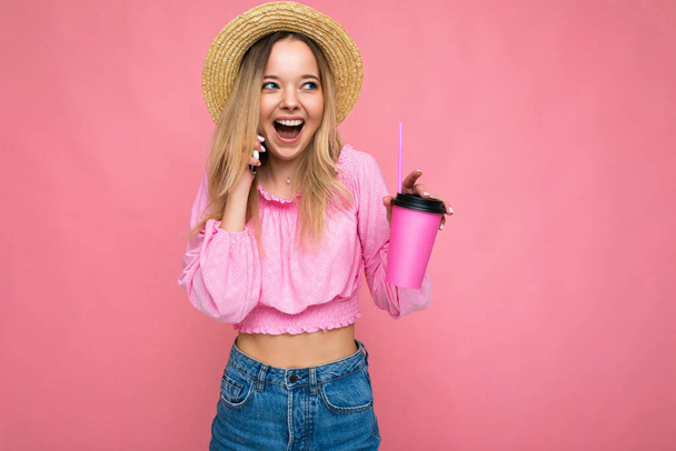 Foto de hermosa mujer rubia emocional positiva que usa blusa rosa y sombrero de paja hablando en el teléfono celular mientras bebe bebidas aisladas en el fondo rosa con espacio para copiar - Foto, Imagen