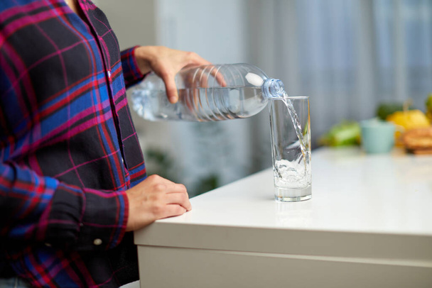 Female hand holding drinking water bottle and pouring water into glass on table on kitchen background. - Foto, immagini
