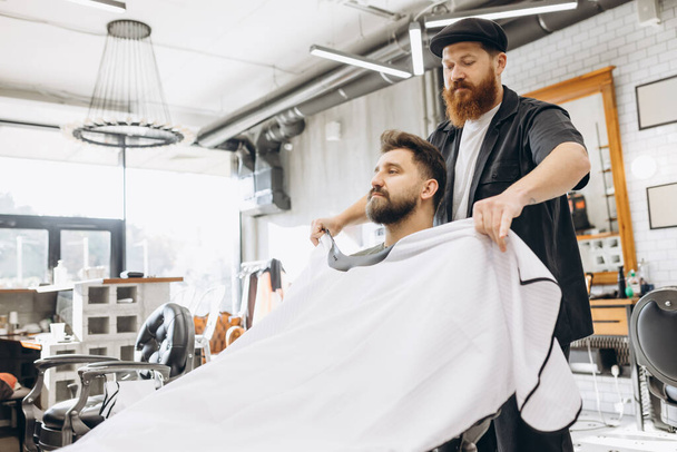 Two handsome men at barbershop. Stylish red-bearded barber and client during haircut process. Beauty, healthcare and male cosmetics concept. - Photo, Image