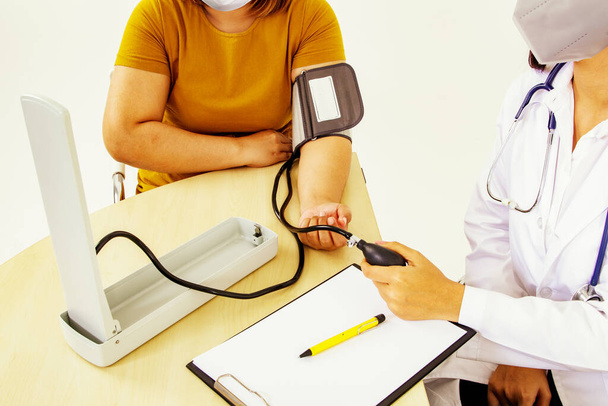 Médico femenino utiliza un monitor de presión arterial en una mujer con presión arterial alta y obesidad a la clínica: Los pacientes y las médicas usan máscaras para prevenir la propagación de gérmenes durante el diagnóstico. - Foto, imagen