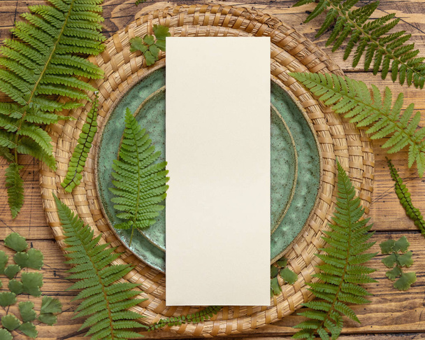 Carta di carta bianca che posa su piatto verde su tavolo di legno marrone con foglie di felce vista dall'alto. Scena di modello tropicale con carta d'invito piatto laici - Foto, immagini