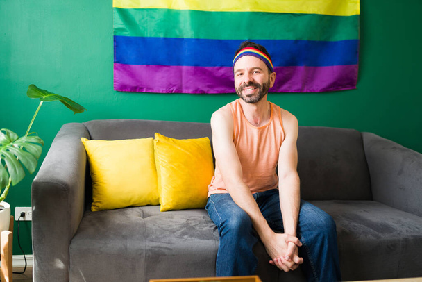 Портрет счастливого гея, улыбающегося и смотрящего в камеру, сидящего дома с радужным флагом позади  - Фото, изображение