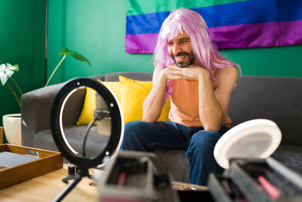 Μου αρέσει να κάνω drag. Queer latin άνθρωπος με μια ροζ περούκα αισθάνεται χαρούμενος και χαμογελώντας, ενώ βάζοντας μακιγιάζ για μια ομορφιά βίντεο φροντιστήριο - Φωτογραφία, εικόνα