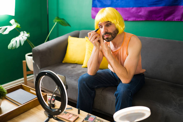 Hermosa persona transgénero con una peluca rubia poniendo delineador de ojos y productos de maquillaje durante un flujo de vídeo en línea - Foto, Imagen