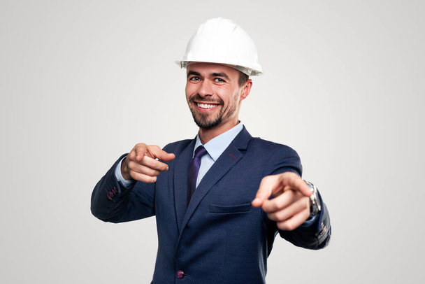 Confident smiling adult bearded male construction manager in formal suit and hardhat pointing at camera while representing job recruitment against white background - Photo, Image