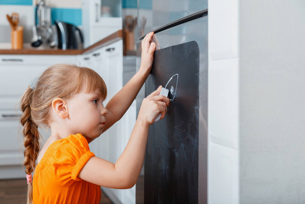 Little caucasian focused child girl draw with chalk on chalk board on refrigerator in kitchen at cozy home interior.  - Фото, изображение