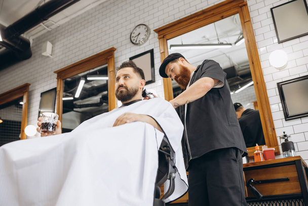 Two handsome men at barbershop. Stylish red-bearded barber and client during haircut process. Beauty, healthcare and male cosmetics concept. - Foto, afbeelding