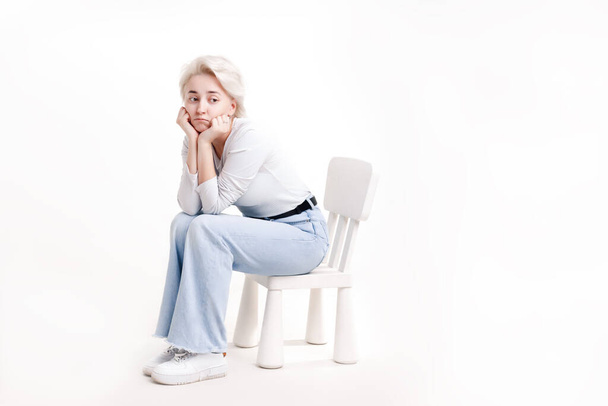 A young blonde girl on a white background sits on a small childrens chair and is sad, in full growth - Photo, Image
