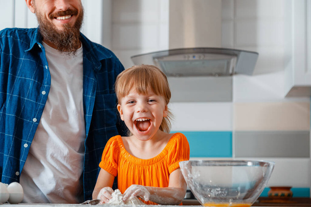 Onnellinen perhe tytär ja isä hauskaa nauraa hymyillä samalla ruoanlaitto ruokaa keittiössä sisustus. Lapsi tyttö tytär vaivaa jauhot taikina lähellä isä ruoanlaitto yhdessä vastaan keittiö sisustus. - Valokuva, kuva