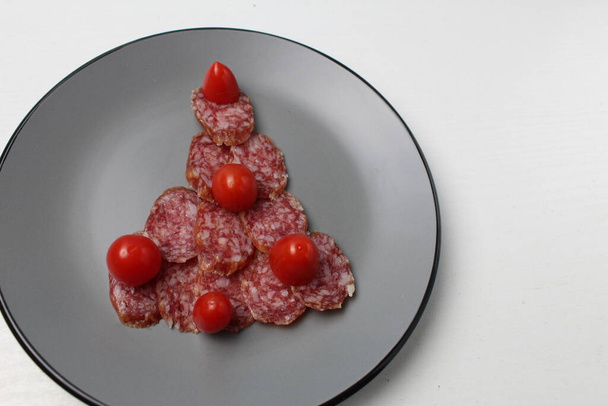 jadalne choinki wykonane z kiełbasy i pomidora wiśniowego leży na szarym talerzu na białym tle światła z miejscem na tekst. - Zdjęcie, obraz