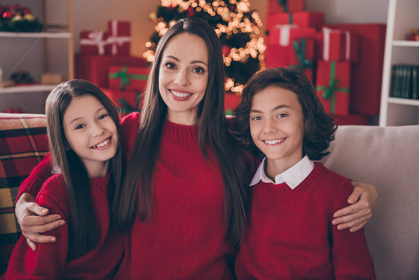 Φωτογραφία από ονειρεμένη γλυκιά μαμά δύο παιδιά ντυμένα κόκκινα πουλόβερ αγκαλιάζει χαμογελώντας γιορτάζει το νέο έτος σε εσωτερικούς χώρους σπίτι δωμάτιο - Φωτογραφία, εικόνα