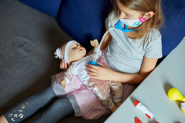 Malá holčička v lékařské masce sedí na pohovce, hraje si na doktora, léčí a poslouchá panenku se stetoskopem v lékařské masce, doma během karantény a pandemie - Fotografie, Obrázek