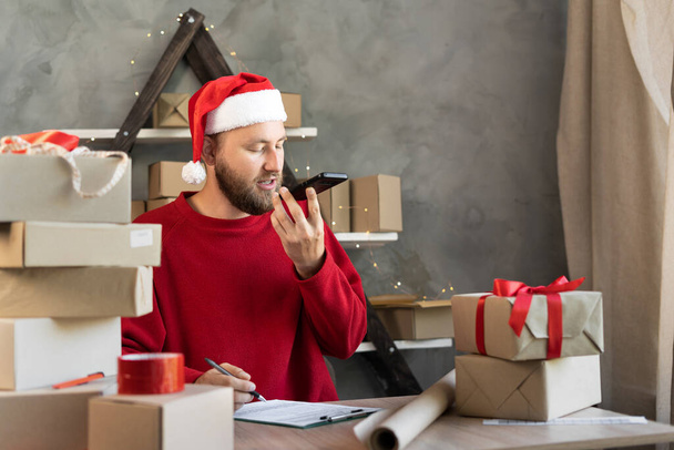 Holidays and Christmas gifts small business delivery service. A man in a Santa Claus hat sitting between boxes with gifts for the new year. Young bearded businessman santa dictates a message to phone - Foto, imagen