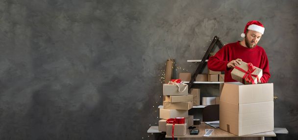 Ένας γενειοφόρος με καπέλο Αϊ Βασίλη δουλεύει σε μια αποθήκη με κουτιά με δώρα και παραγγελίες από ένα ηλεκτρονικό κατάστημα για τα Χριστούγεννα. Ο ιδιοκτήτης των μικρών επιχειρήσεων πουλάει Καλή Χρονιά. Διακοπές παράδοσης. banner - Φωτογραφία, εικόνα