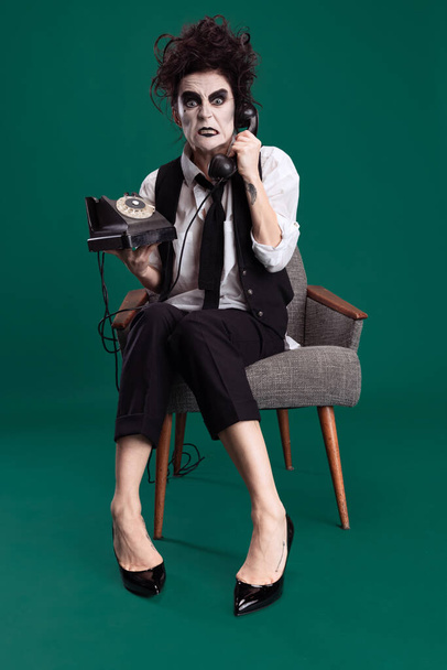Mujer espeluznante, espeluznante en traje femenino blanco y negro con maquillaje loco y cabello desordenado sosteniendo teléfono vintage aislado sobre fondo verde - Foto, imagen
