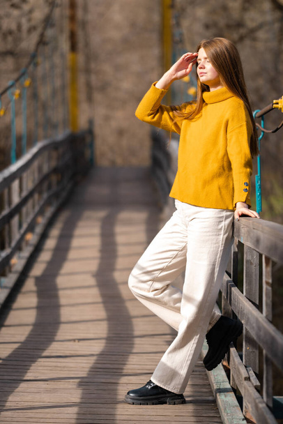 Рудоволоса дівчина стоїть на дерев'яному мосту в помаранчевому светрі і дивиться на осіннє сонце. Розслаблена молода жінка гуляє в лісі
. - Фото, зображення