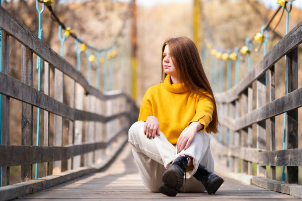 Рыжая девушка сидит на деревянном мосту в оранжевом свитере и смотрит на осеннее солнце. Расслабленная молодая женщина ходит по лесу. - Фото, изображение