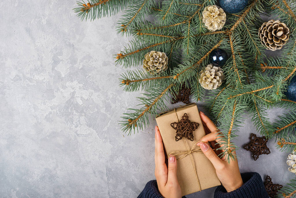 Female hands hold a gift box on a gray background. Composition with cones and spruce branches.Festive background for the holidays: Christmas, New Year. Flat lay. - Fotoğraf, Görsel