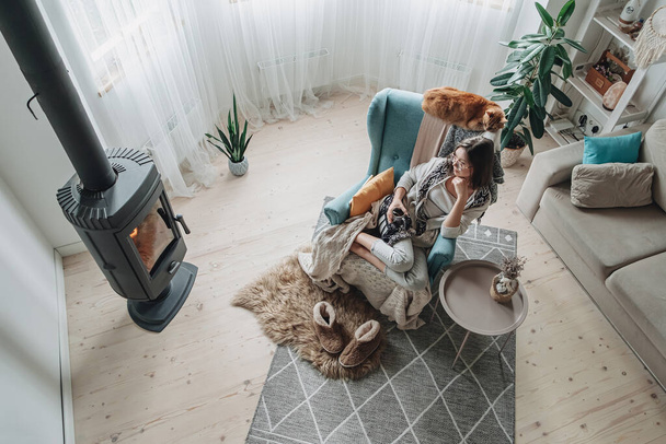 Young woman in a cozy armchair and a plaid sitting by the fireplace with a domestic cat and drinking tea at home - Foto, Bild