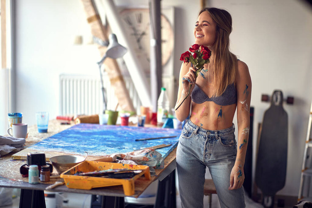 A young sexy and attractive female artist in a relaxed atmosphere in her studio is holding roses she got and thinking. Art, painting, studio - Foto, afbeelding