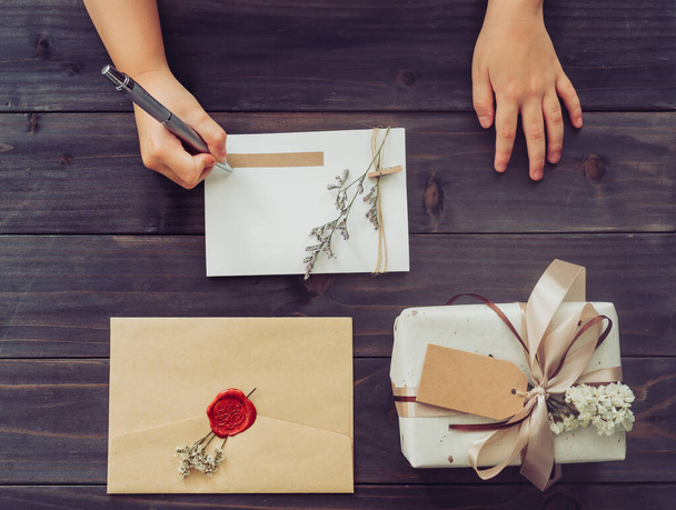 Boven van de hand meisje schrijven kaart en ambachtelijke geschenkdoos op houten tafel achtergrond  - Foto, afbeelding
