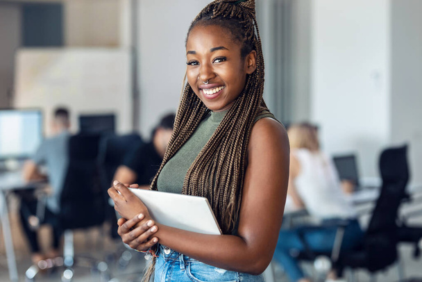 Porträt einer lächelnden jungen Unternehmerin, die ihr digitales Tablet benutzt, während sie an einem Coworking-Platz vor der Kamera steht. - Foto, Bild
