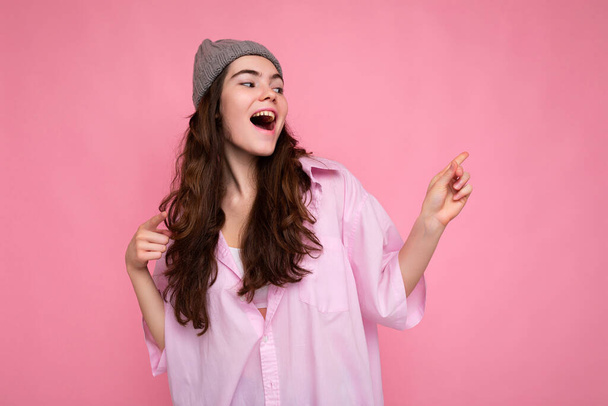 Красива щаслива щаслива молода кучерява брюнетка в рожевій сорочці та сірому капелюсі ізольована на рожевому фоні з місцем для тексту
 - Фото, зображення