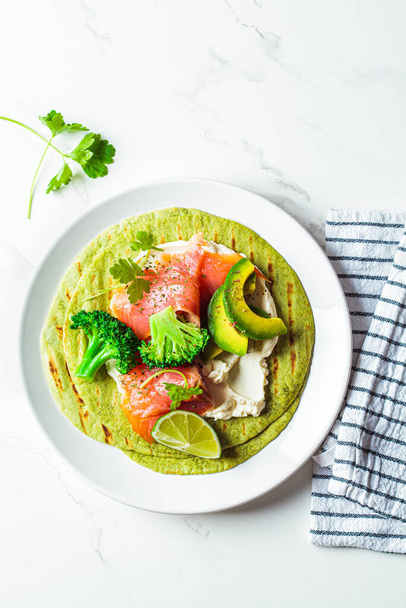 Green tortilla with broccoli, avocado, cream cheese and salted salmon on a white marble background. Vegetarian fast food. - Photo, Image