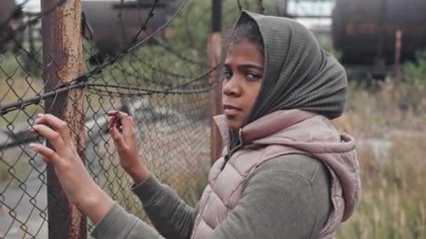 Medium close-up portrait of 11-year-old abandoned refugee girl of African-American ethnicity standing outdoors with hands on barbed-wire fence looking at camera - Footage, Video