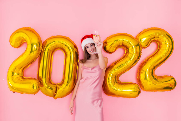 Excited girl raises up glass of champagne in Santas hat. 2022 golden air balloons New Year concept  - 写真・画像