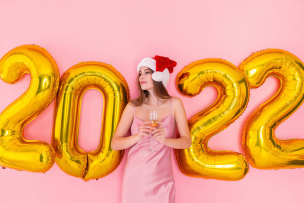 Excited woman holds glass of champagne in Santas hat looks away. golden air balloons 2022 New Year - Zdjęcie, obraz