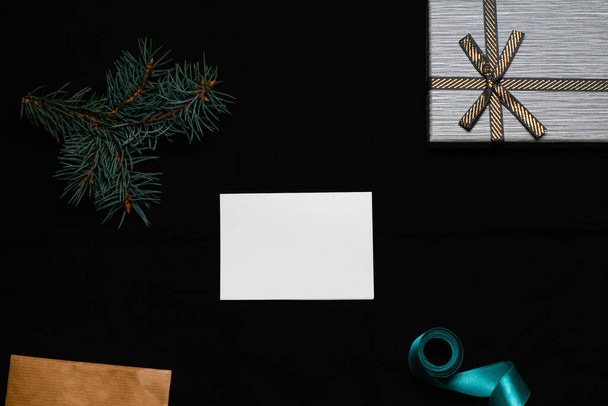 Defocus blank white card with spruce tree branch, gift bow, craft envelope and blue ribbon on black background. Frame mockup, mock up with empty sheet of paper. Xmas holiday concept. Out of focus. - Photo, image