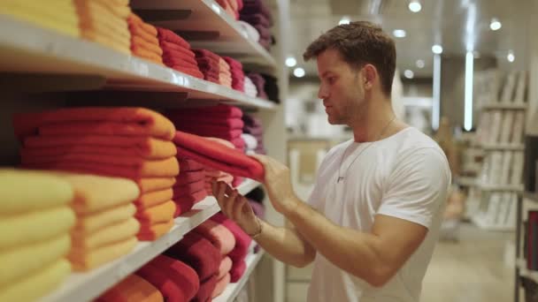 Молодой бородатый мужчина выбирает яркие красочные полотенца с полки во время посещения отдела товаров для дома в современном магазине - Кадры, видео