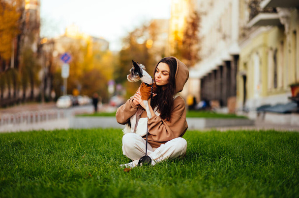Attractive lady is sitting on a green lawn with her cute small dog - yorkie. Blurred background, city street. - Photo, Image