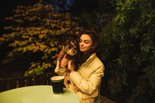Atraktivní běloška sedí na terase kavárny se svým roztomilým yorkteriérem, večer pije kávu. - Fotografie, Obrázek