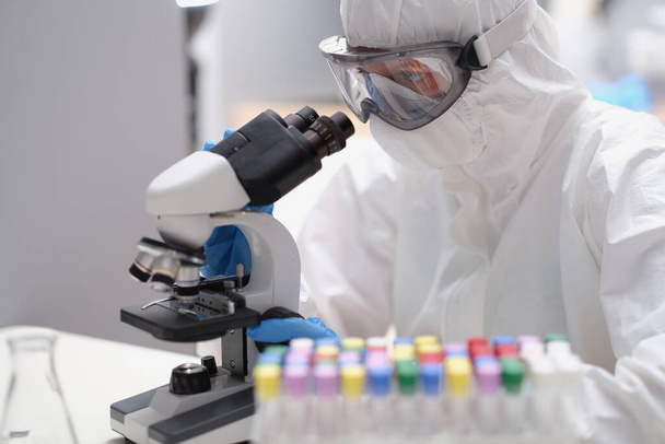 Tudós védőruhában és maszkban mikroszkóppal néz - Fotó, kép
