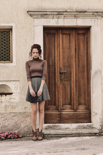 Retrato vintage tonificado de una joven mujer esperando frente a una vieja puerta de madera al lado de la carretera mirando al suelo - Foto, imagen