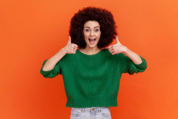 Amazed positive woman with Afro hairstyle wearing green casual style sweater standing showing thumb up with both hands, recommend content. Indoor studio shot isolated on orange background. - Фото, изображение