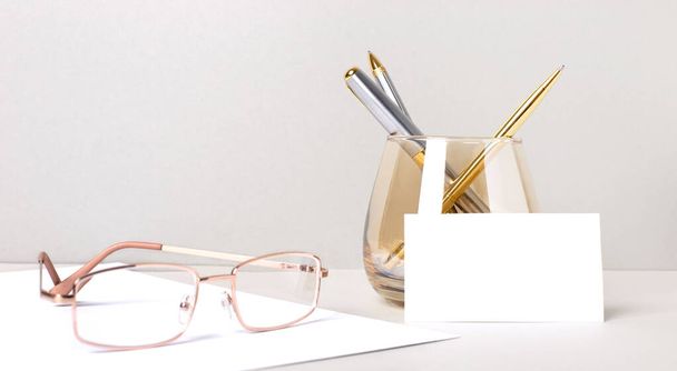 Work desk with gold glasses, pens in a glass and white blank card with place to insert text. Business template. Home Office. Workplace close-up. - Foto, Imagem