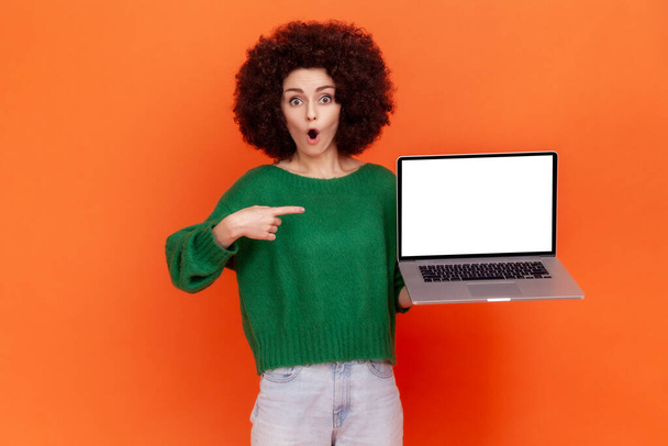 Shocked woman with Afro hairstyle wearing green casual style sweater pointing at white empty display of her portable computer, copy space. Indoor studio shot isolated on orange background. - Foto, Imagen