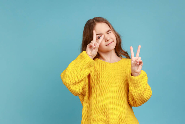 Portrait of little girl showing v sign with double fingers, gesturing victory, celebrating success, wearing yellow casual style sweater. Indoor studio shot isolated on blue background. - Fotografie, Obrázek