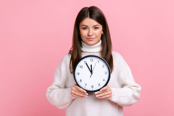 Portrait of serious female holding big wall clock, looking at camera with pleasant smile, time to go, wearing white casual style sweater. Indoor studio shot isolated on pink background. - 写真・画像